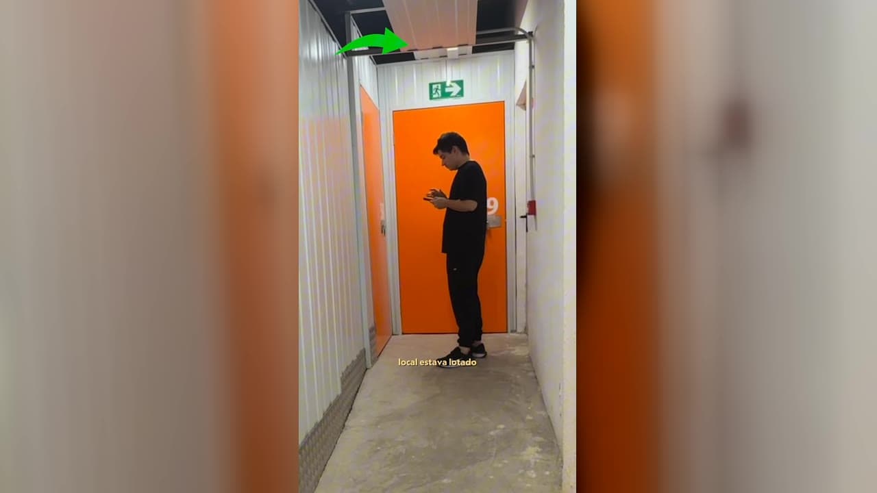 Red no corredor do depósito, olhando o celular para localizar o rastreador da mala, com uma seta apontando para o teto em grade